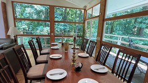 A dining area with a long wooden table set for six, surrounded by wooden chairs. Large windows offer a view of a forested area. - 5-Bedroom 6000SF wood and glass retreat in Atlanta, surrounded by greenery.   (Atlanta)