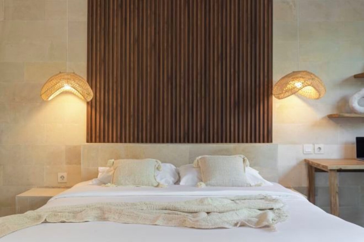 Habitación de hotel con una cama grande, dos lámparas de noche, un cabecero de listones de madera y una pequeña mesa con una silla.