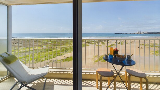 Ein Balkon mit Blick auf den Strand, ein Tisch mit Flasche und Gläsern sowie ein Stuhl.