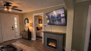 A living room with a fireplace, a television mounted above it, and a cityscape painting on the wall. - Hot Tub+Arcade / Fire Pit Retreat Near Monon Trail (Indianapolis)