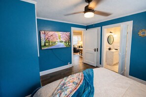Une chambre à coucher avec un lit, un ventilateur de plafond, un tableau représentant une route bordée d’arbres, et une salle de bain visible à travers une porte ouverte.