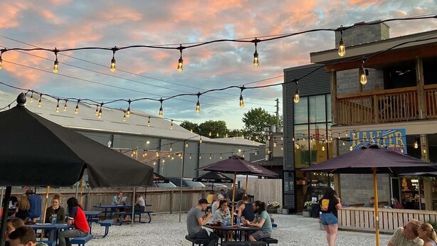 An outdoor seating area with tables and umbrellas, a building with a sign reading "HALTER," and string lights overhead. - Hot Tub+Arcade / Fire Pit Retreat Near Monon Trail (Indianapolis)