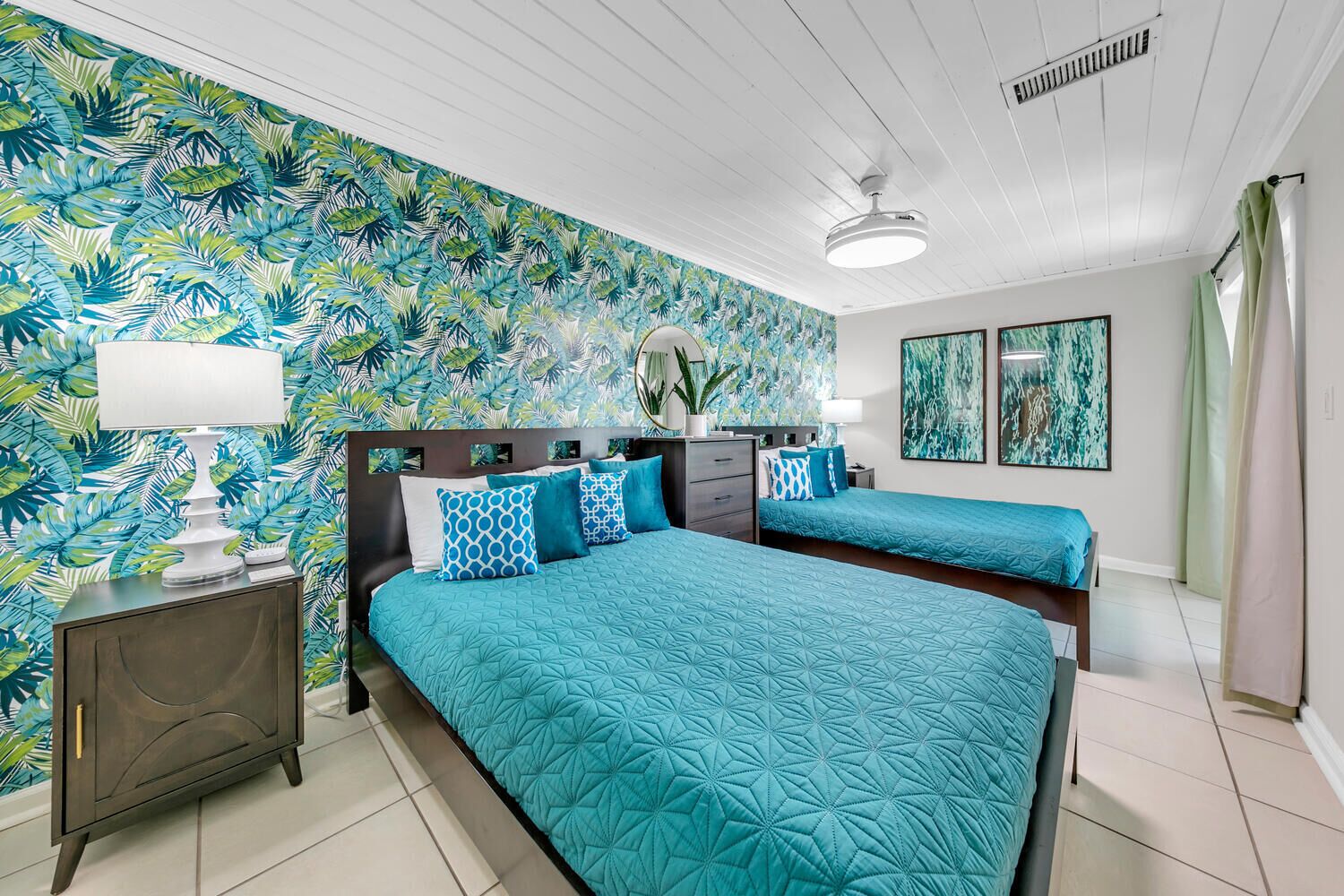 Un dormitorio con una cama grande, una cama más pequeña, una mesita de noche con lámpara, un aparador y un papel tapiz con hojas tropicales.