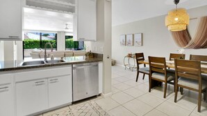 A modern kitchen with white cabinets, a stainless steel dishwasher, and a dining area with wooden chairs and a table. - The Springs: Luxury Resort Living, Palm Springs (Palm Springs)