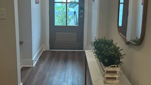 Un couloir avec un plancher en bois, une porte dotée d’une fenêtre, un miroir accroché au mur et une plante posée sur une table blanche.