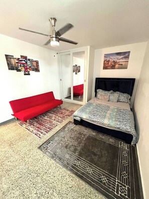 A bedroom with a bed, a red sofa, a ceiling fan, and a view of a cityscape through a window. - Stylish Remodeled 3BR With Pool,parking Near Airport & Beaches (Carolina)
