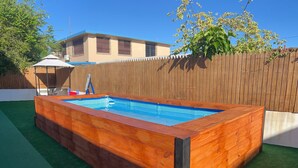 Una vasca idromassaggio in legno rettangolare con interni in piastrelle blu, circondata da una recinzione in legno e prato sintetico verde.