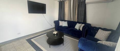 A living room with a dark blue sofa, a small round coffee table, a ceiling fan, and a flat-screen TV mounted on the wall.