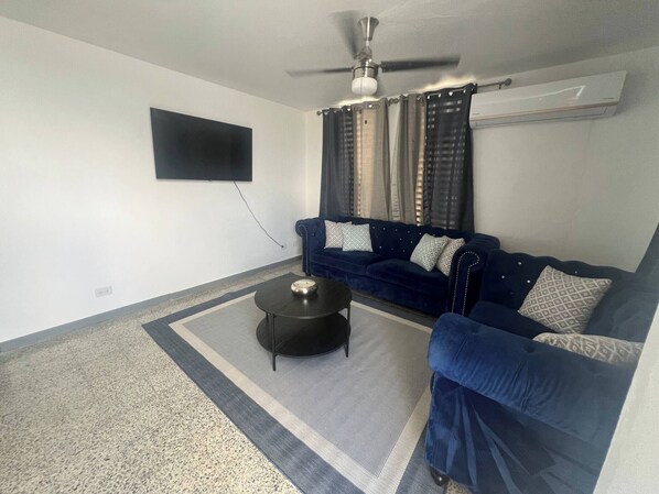 Un salon avec un canapé bleu foncé, une petite table basse ronde, un ventilateur de plafond et un téléviseur à écran plat fixé au mur.