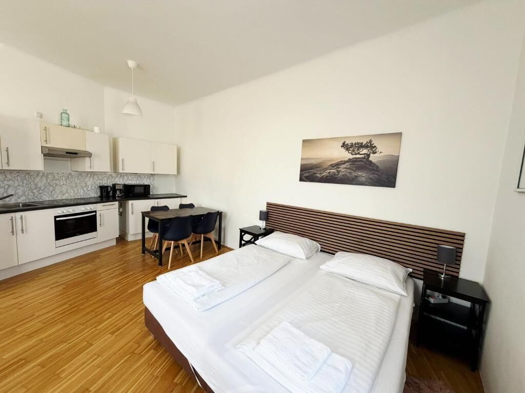A modern hotel room with a wooden floor, a bed with white linens, a small desk with a chair, a kitchenette, and a painting of a tree on the wall.