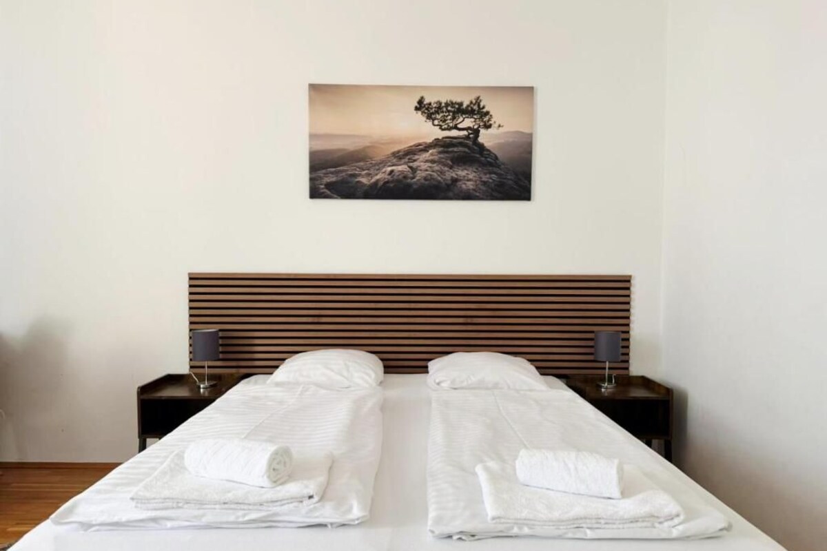 A double bed with white linens and towels, a wooden headboard, and a framed picture of a tree on a mountain.