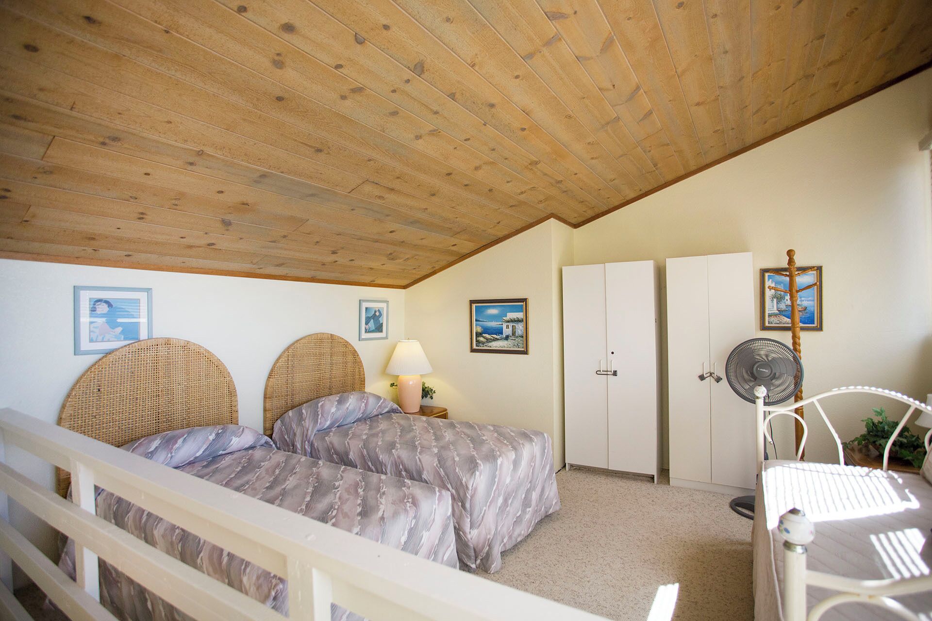 A bedroom with a wooden ceiling, two large wicker headboards, a bed with patterned bedding, a bedside table with a lamp, a wardrobe, a standing fan, and framed pictures on the wall.