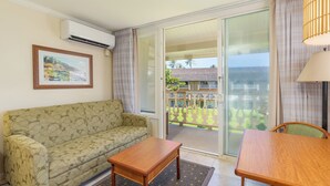 A living room with a sofa, a wooden table, and a view of a balcony with a railing and greenery. - Studio w/Ocean Views - Islander On The Beach 322 (Kapa'a)