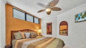 A bedroom with a large bed, a ceiling fan, a window with blinds, and a painting of palm trees.