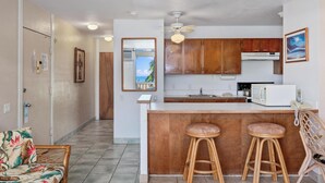 A compact kitchen with wooden cabinets, a white countertop, and two stools.