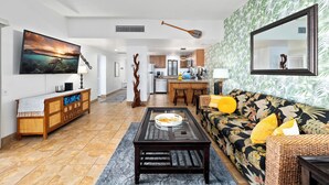 Una sala de estar con sofá, mesa de centro y televisor en la pared. Se ve una cocina al fondo.