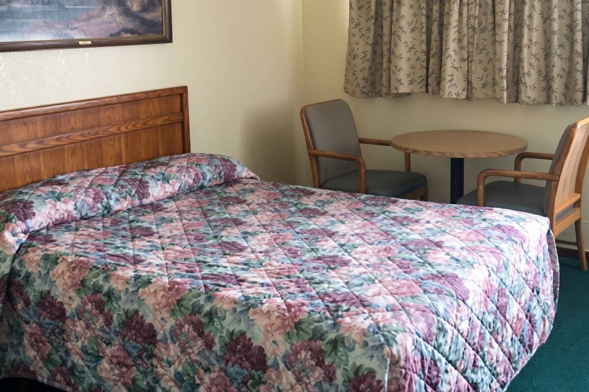 Un lit avec une couette à motifs floraux, une tête de lit en bois, une petite table ronde avec deux chaises et une fenêtre ornée de rideaux à motifs.
