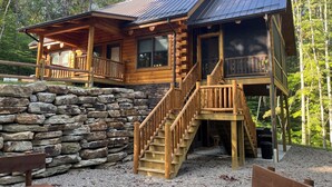 Eine Holzhütte mit Steinfundament, einer Holzdielen-Terrasse und einer Treppe, die zu einem Balkon führt.