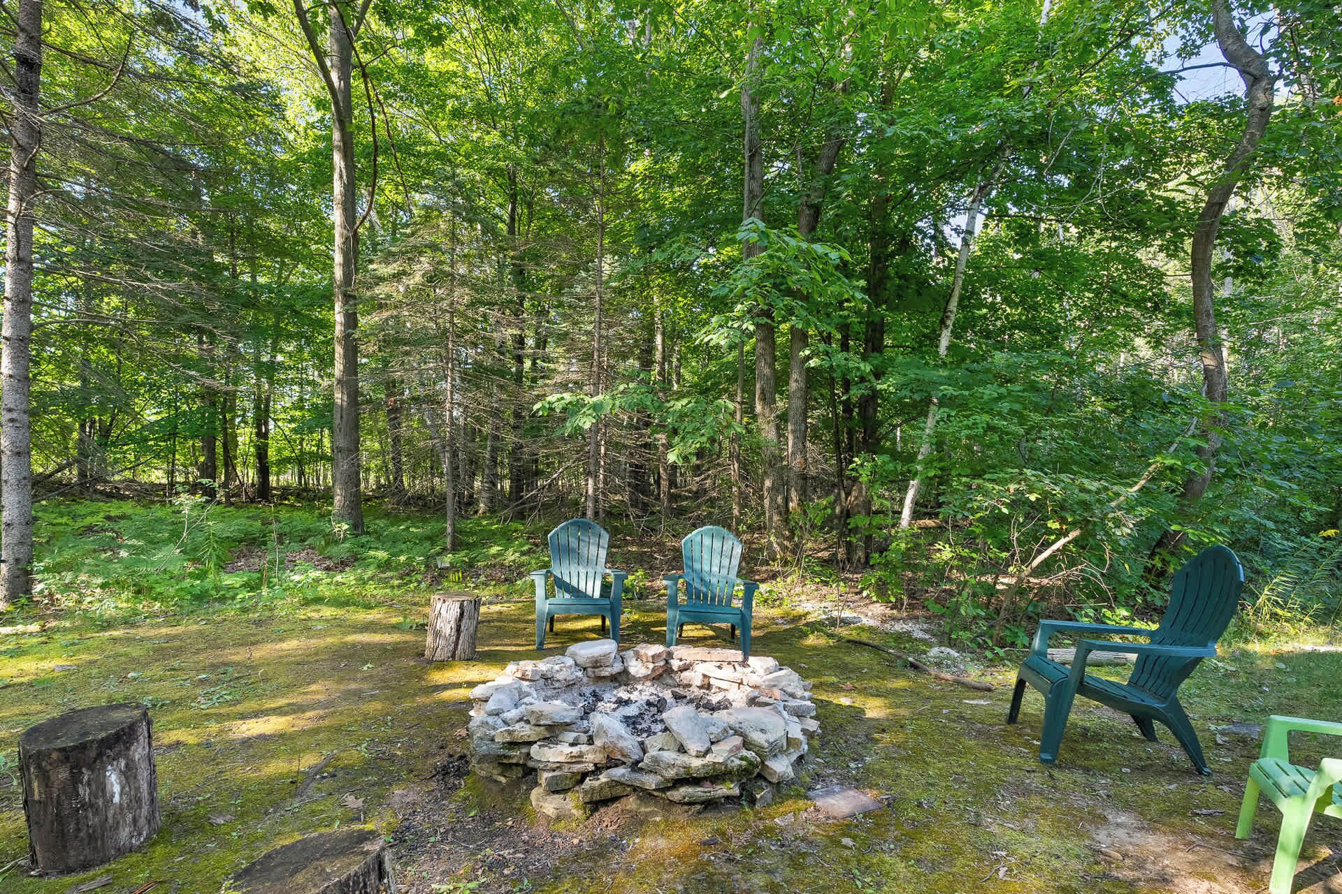 A forested area with a stone fire pit and three green Adirondack chairs.