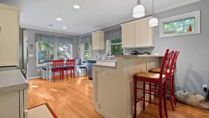 A kitchen with wooden flooring, a dining area with red chairs, and a window with a view of greenery. - Blue Bear Cottage: Serene Fish Creek Escape (Fish Creek)