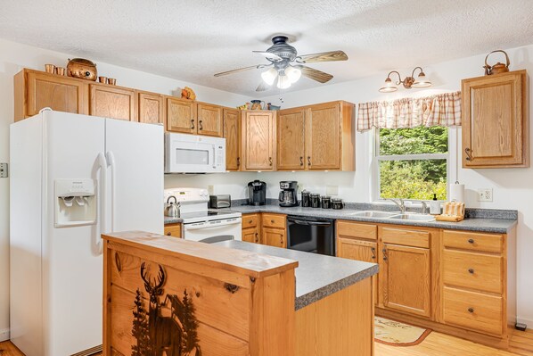 A kitchen with wooden cabinets, a white refrigerator, a microwave, a coffee maker, and a window with a view of greenery. - Hot Tub, Fire Pit & Mountain Views + Biltmore Pass | Cozy One-Level Cabin (Waynesville)