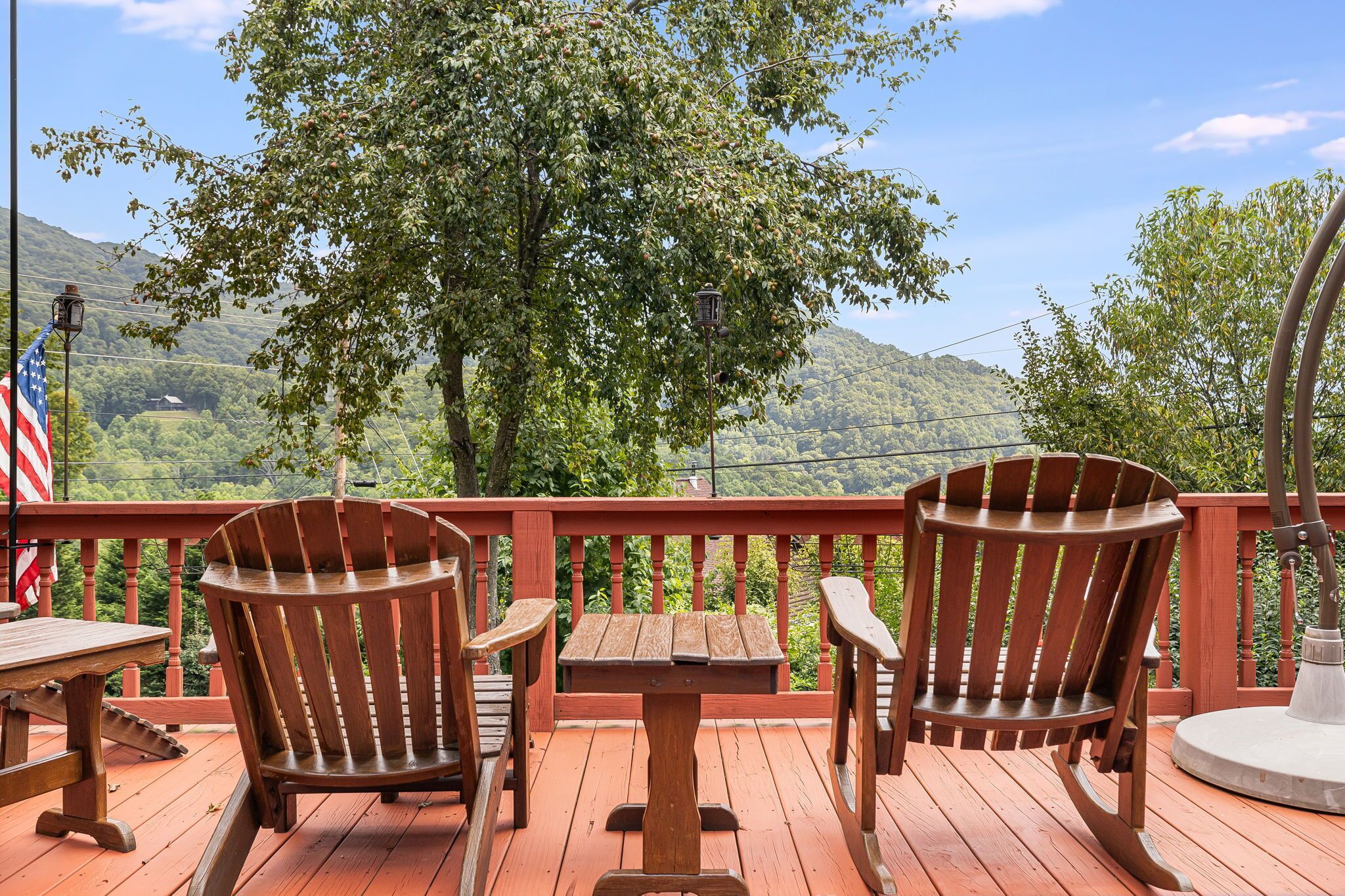A wooden deck with two Adirondack chairs and a small table, overlooking a lush green landscape with hills.