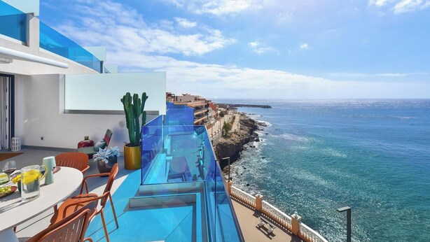 Une terrasse extérieure moderne, dotée d’une barrière en verre offrant une vue sur la mer, comprend un espace repas avec des chaises et une table, un cactus et une vue sur la côte.