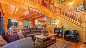 A cozy log cabin interior with a wooden staircase, a sofa set, a dining area, and a fireplace.