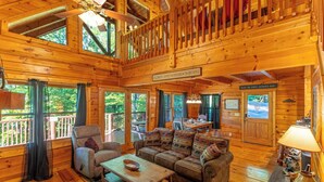 A spacious wooden cabin with a high ceiling, a cozy living room with a sofa set, a dining area, and a balcony with a railing.