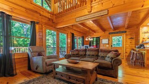 A cozy log cabin interior with a wooden ceiling, a sofa set, a chair, a coffee table, and a view of the forest through large windows.