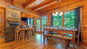 A cozy cabin interior with a stone fireplace, wooden dining table, and a large window offering a view of greenery.