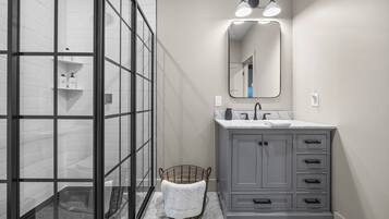 A bathroom with a glass shower enclosure, a gray vanity with a mirror, and a wicker basket with a white towel.