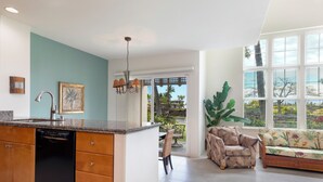 A spacious kitchen with a dishwasher, a dining area with a table and chairs, a sofa with a tropical print, and large windows offering a view of the outdoors. - Includes Beach Gear!  2404 Waikoloa Colony Villas (Waikoloa Beach Resort)