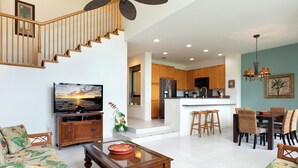 A spacious living room with a sofa, coffee table, and a TV. A kitchen with wooden cabinets and a dining area with chairs is visible in the background. - Includes Beach Gear!  2404 Waikoloa Colony Villas (Waikoloa Beach Resort)