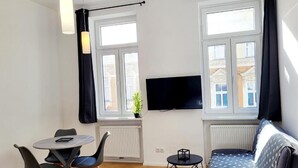 Una sala de estar moderna con un sofá, una mesa de comedor con sillas, un televisor montado en la pared y ventanas amplias con cortinas.