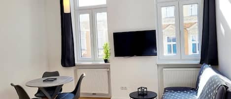 A modern living room with a sofa, a dining table with chairs, a TV mounted on the wall, and large windows with curtains.