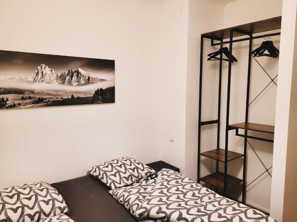 A small room with a bed, a black and white patterned bedspread, a wall-mounted shelf with hangers, and a framed picture of a mountain landscape.