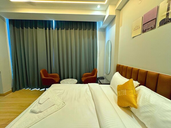 A hotel room with a bed, two chairs, a small table, a mirror, and a window with curtains. - Lime Tree Hotel Sector 51 Gurgaon (Gurugram)