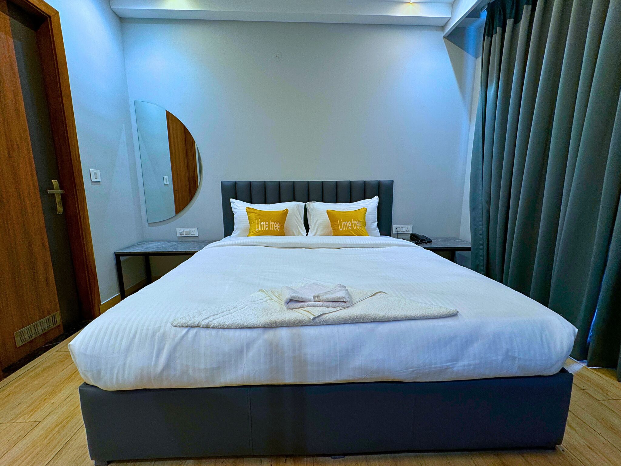 Una cama bien tendida con ropa blanca y dos almohadas, una mesita de noche con un teléfono, y un espejo en la pared.