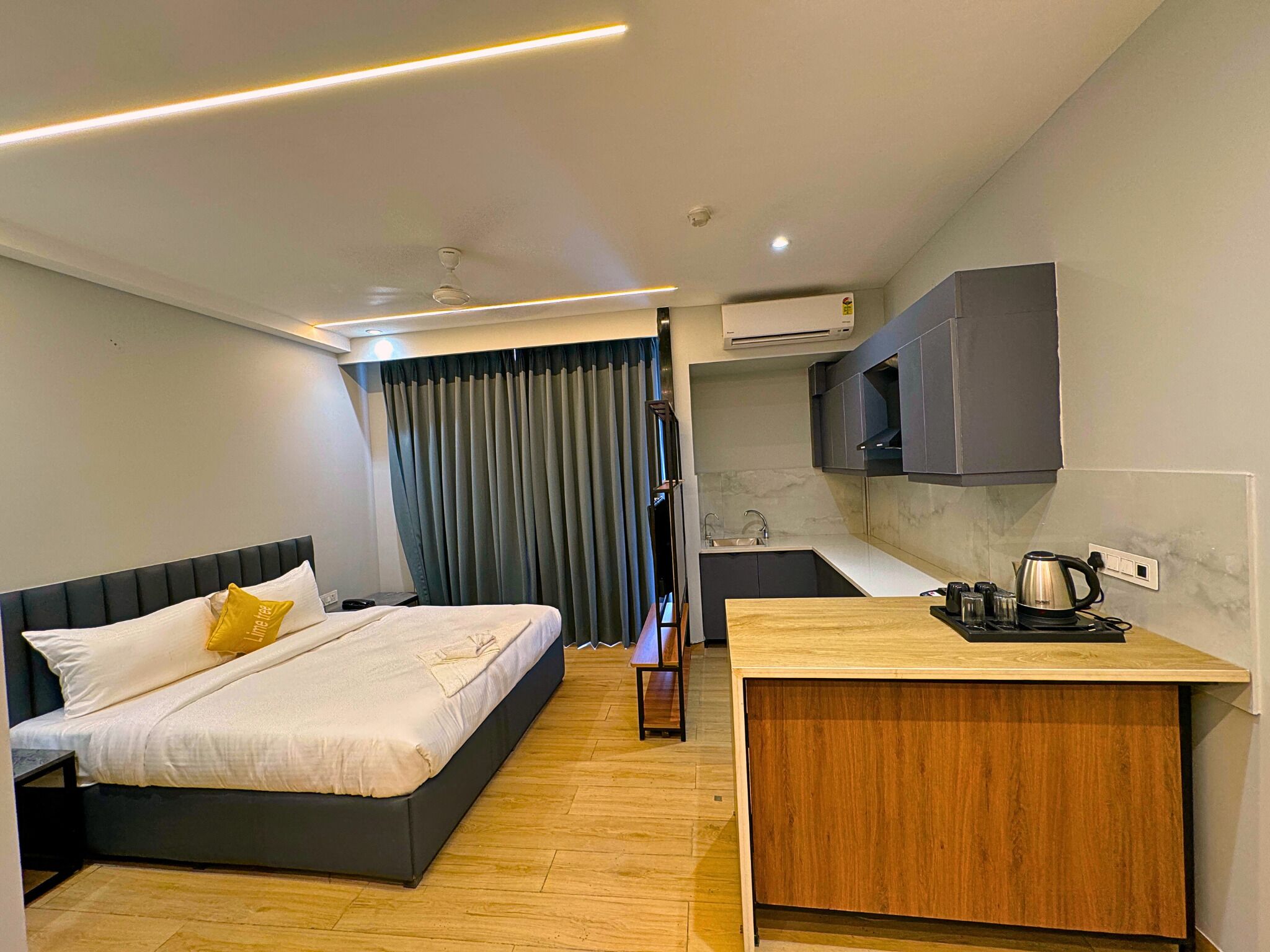 Ein modernes Hotelzimmer mit Bett, einer kleinen Küchenzeile und einem durch eine Glastür einsehbaren Badezimmer.