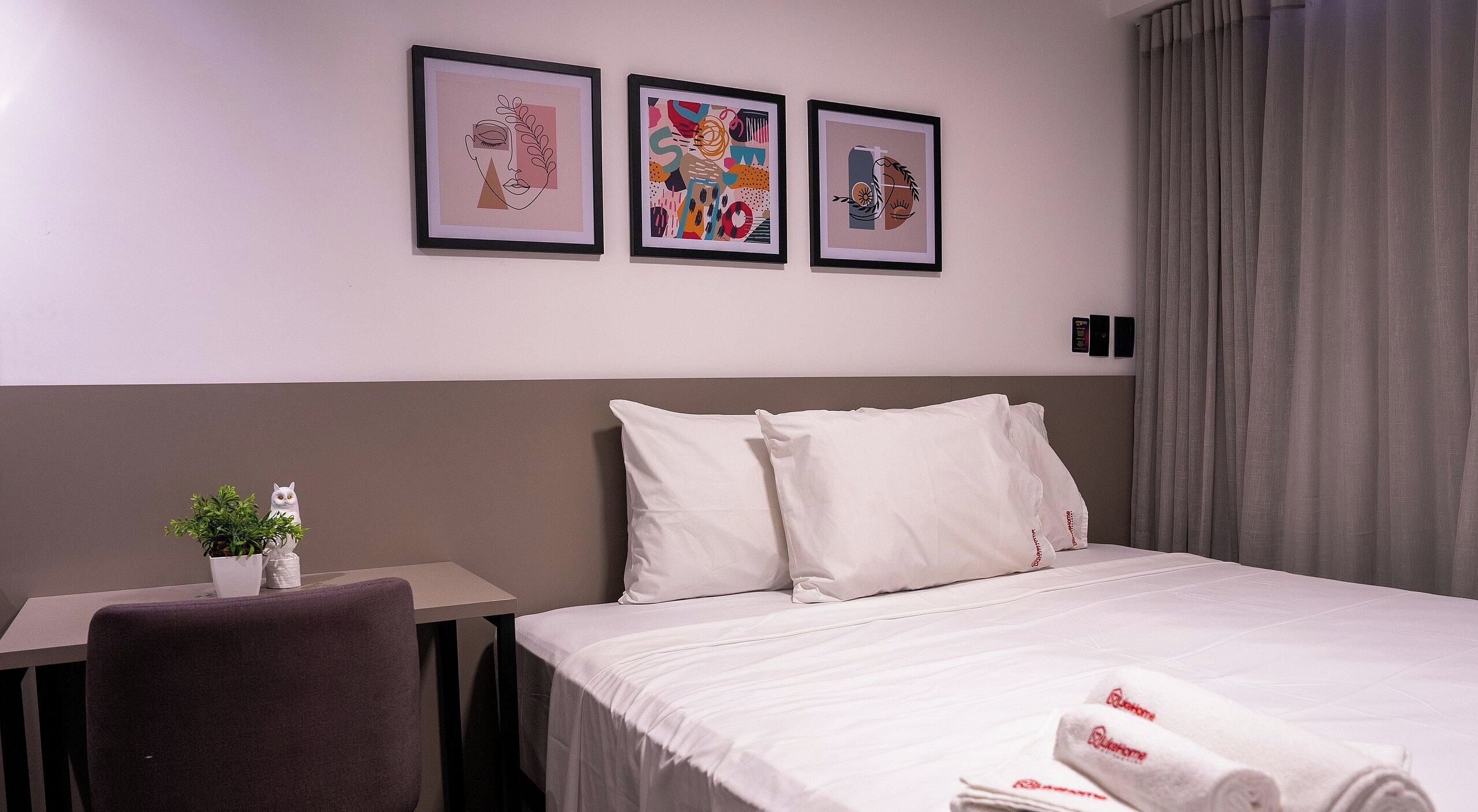 A hotel room with a bed, a desk, and three framed artworks on the wall.