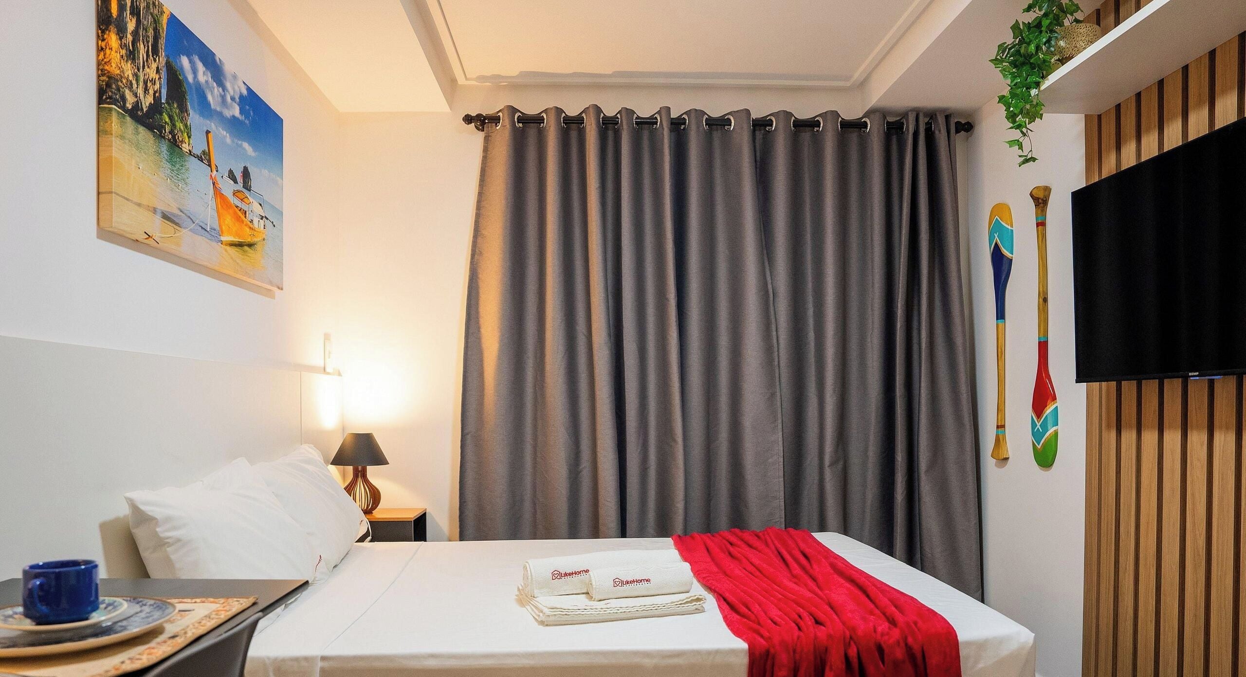 A hotel room with a bed, a painting, a curtain, a TV, and a plant.