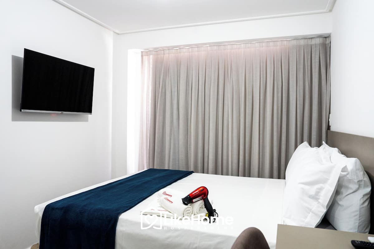 A hotel room with a bed, a TV mounted on the wall, a hairdryer, and a window with curtains.