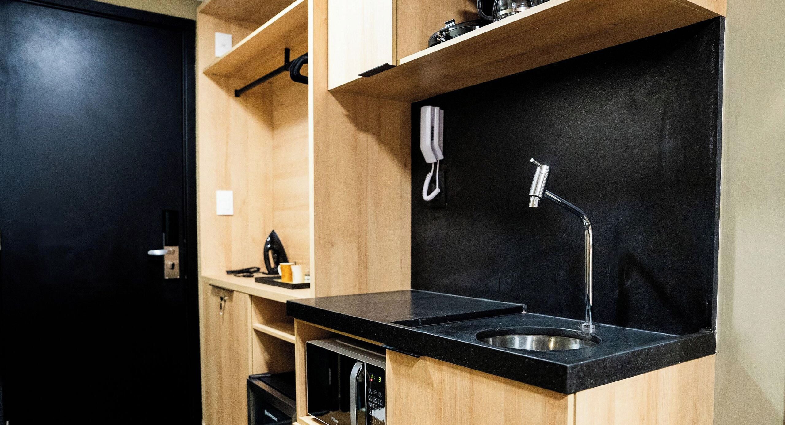 Una kitchenette compacta con cubierta negra, un lavamanos de acero inoxidable, un teléfono blanco y un grifo negro fijado a la pared.