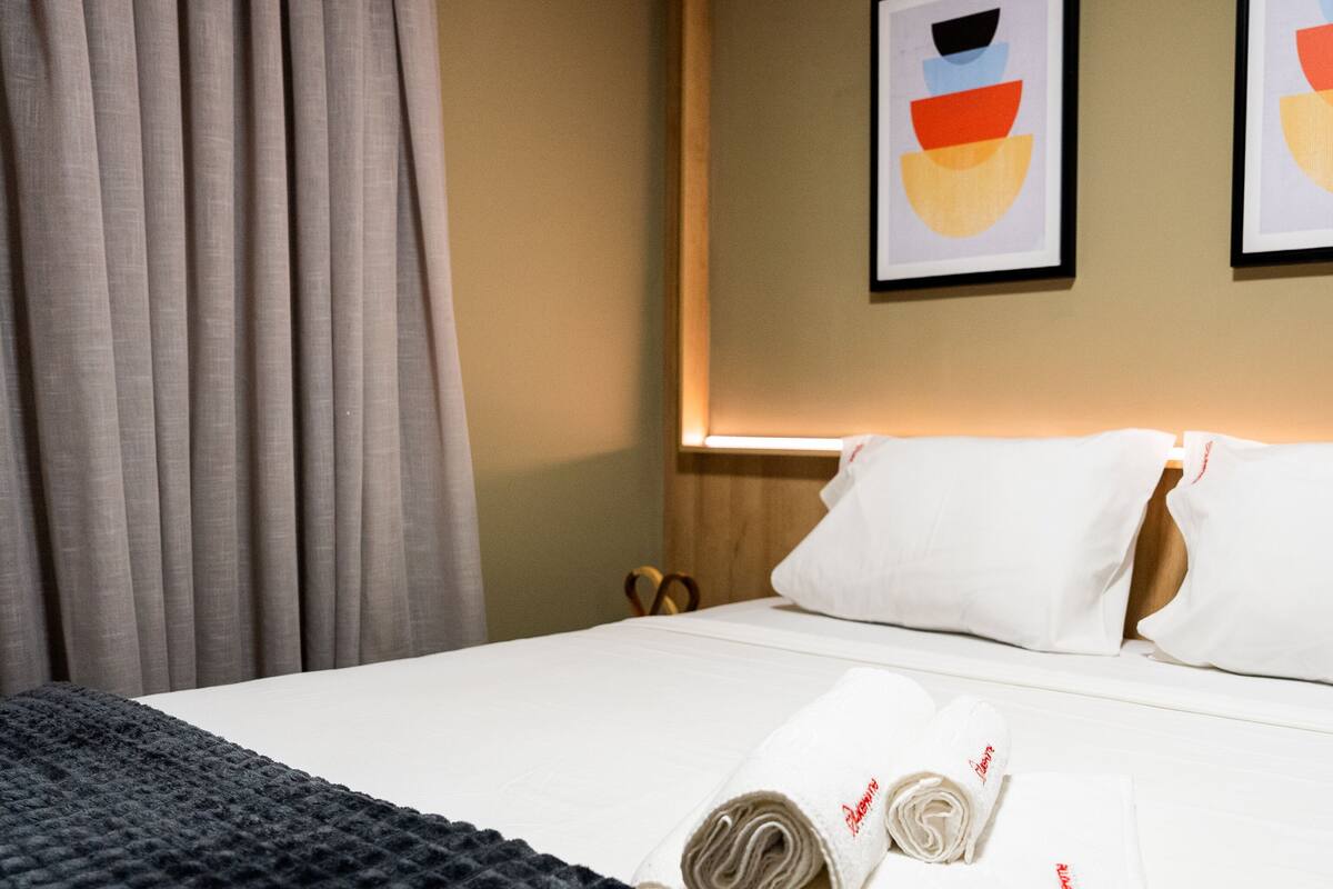 A hotel room with a neatly made bed, white pillows, and a rolled towel on the bed. There are two framed artworks on the wall above the bed.