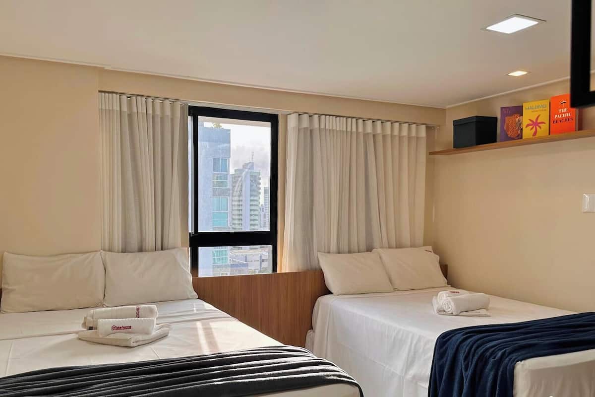 Ein Hotelzimmer mit zwei Betten, einem Fenster mit Vorhängen, Blick auf Gebäude und einem Regal mit Büchern.