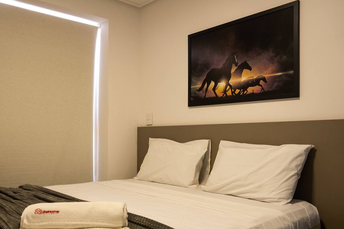A hotel room with a bed, a blanket, pillows, a painting of horses, and a window with blinds.