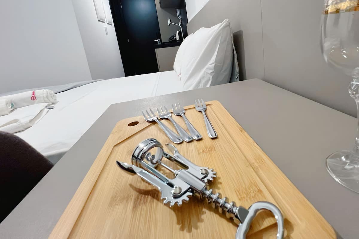 A hotel room with a wooden board displaying a bottle opener, a wine glass, and a set of forks and knives.