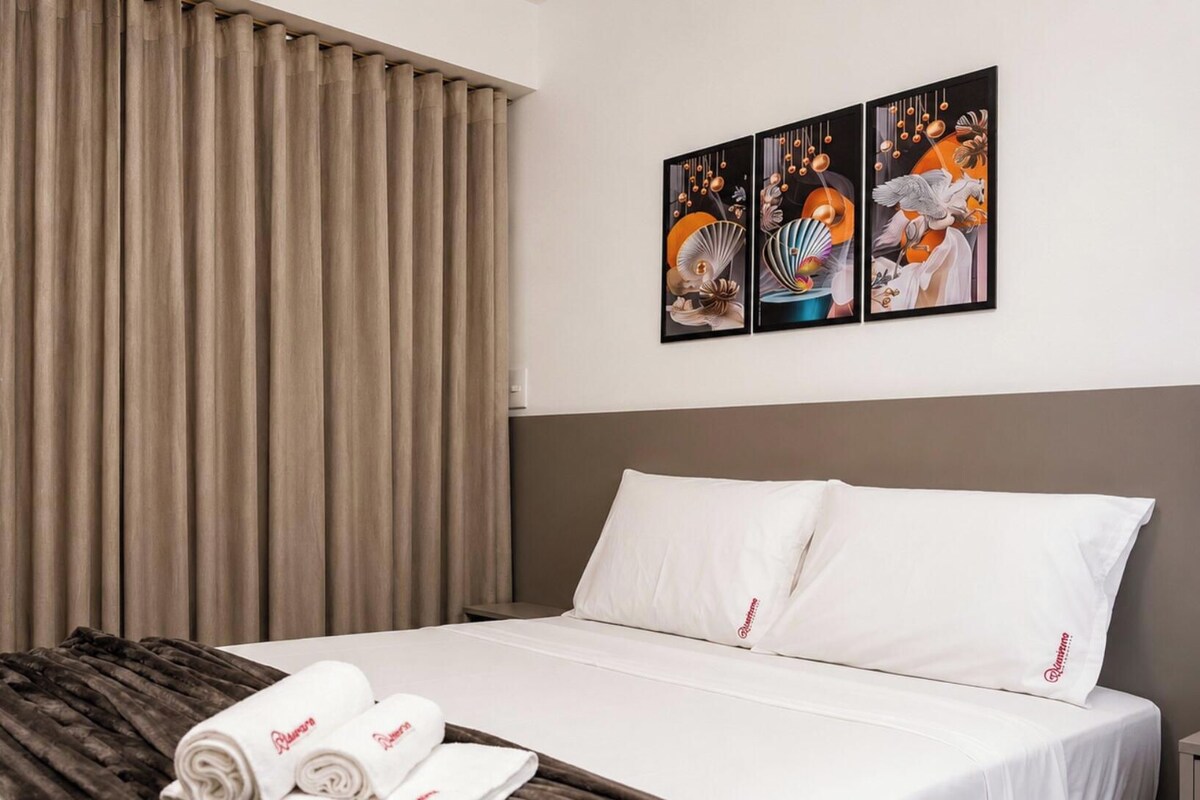 A hotel room with a bed, a set of curtains, a wall-mounted artwork, and a rolled towel on the bed.