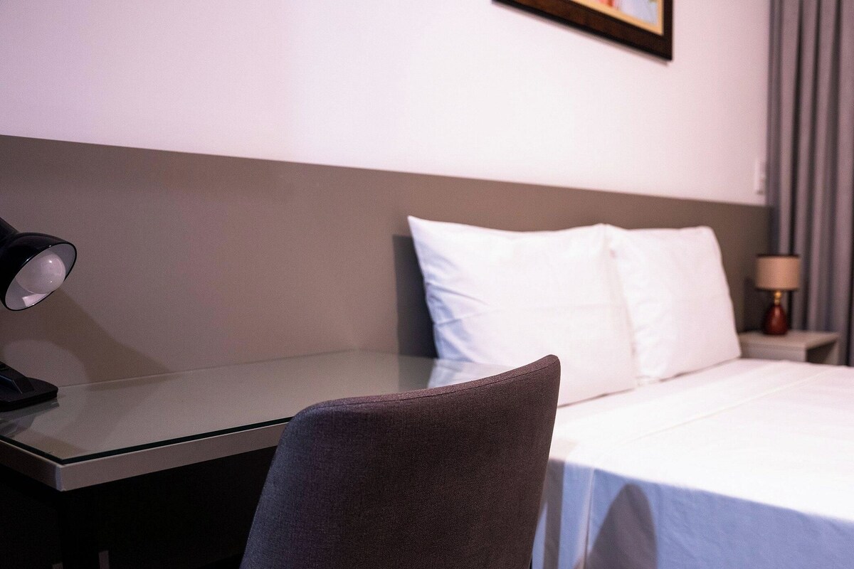 A hotel room with a bed, a desk, a chair, a lamp, and a nightstand.